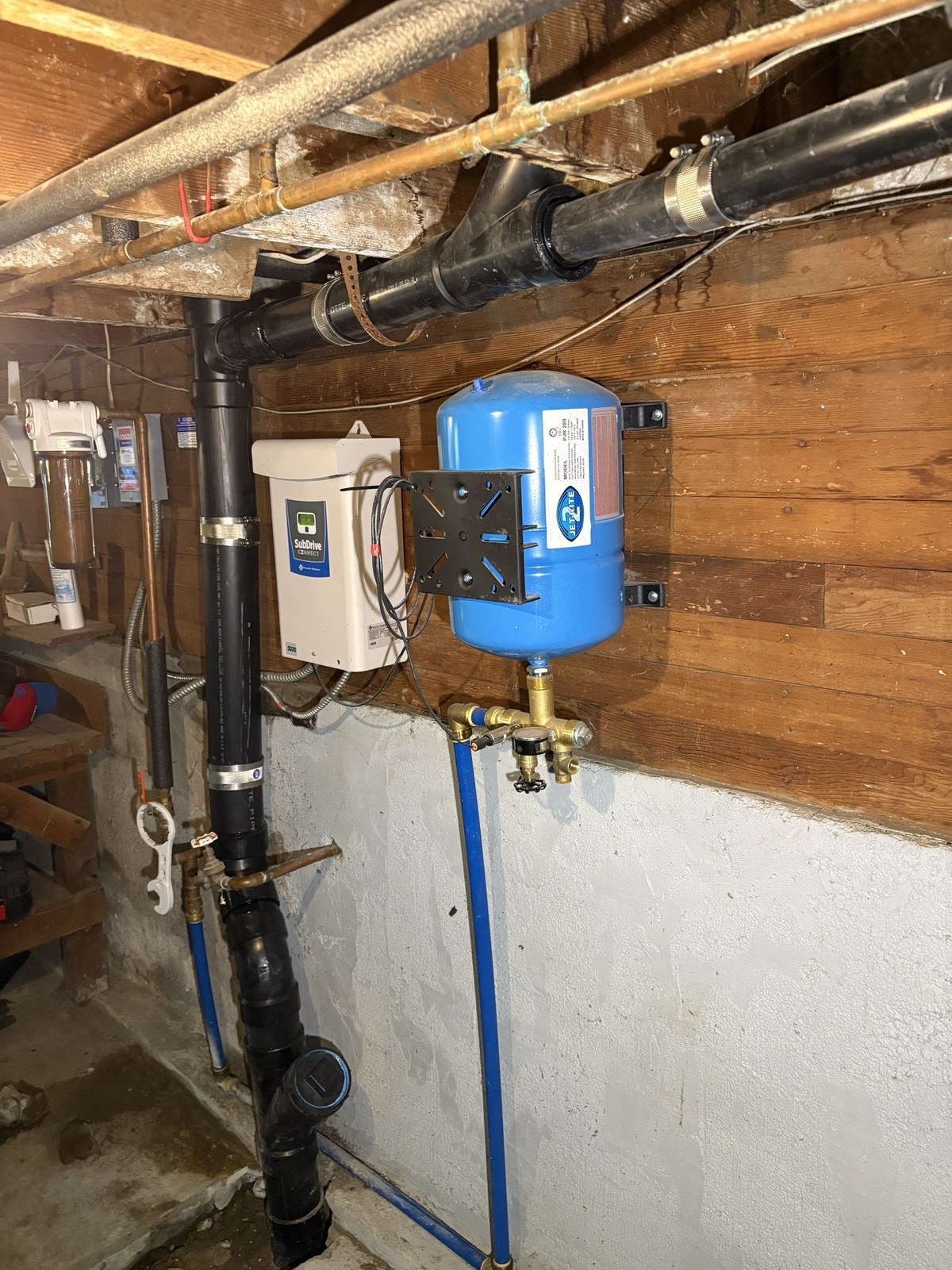 The same basement corner with new black ABS drain pipe and a cleanout fitting dropping into the floor, the blue pressure tank and pump controller still in place