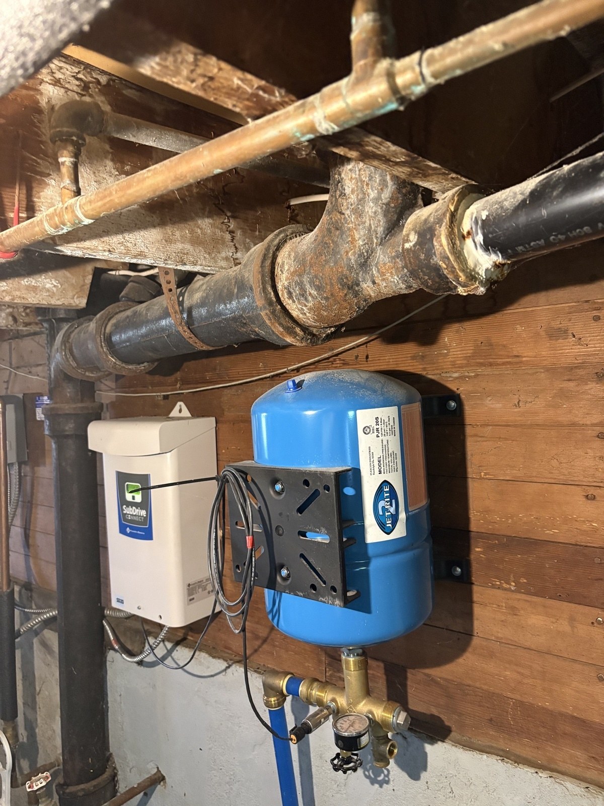 Old black cast-iron drain pipe with crusted, rust-stained joints running across a basement ceiling above a blue well pressure tank and a white pump controller