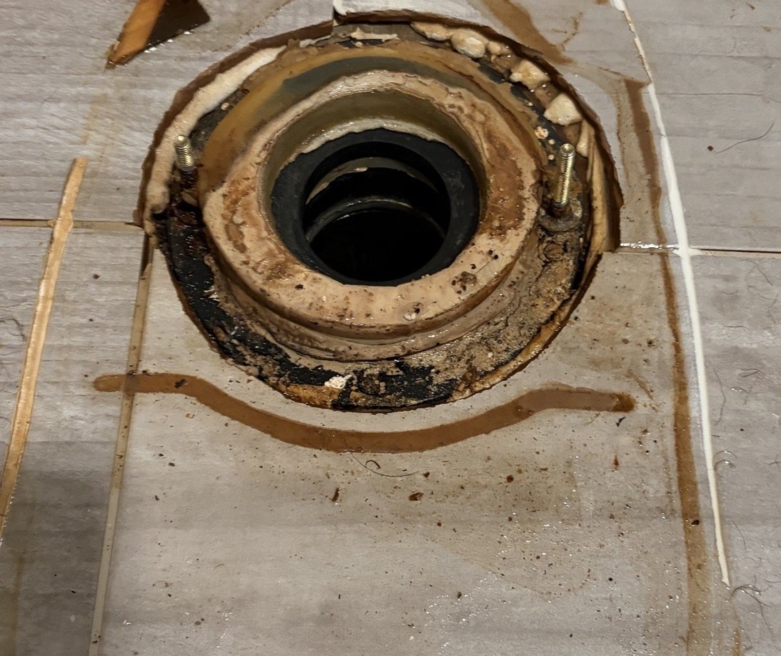 A toilet flange on a tiled bathroom floor after the toilet was lifted, with the old wax seal crushed and stained around the drain opening and water marks on the tile
