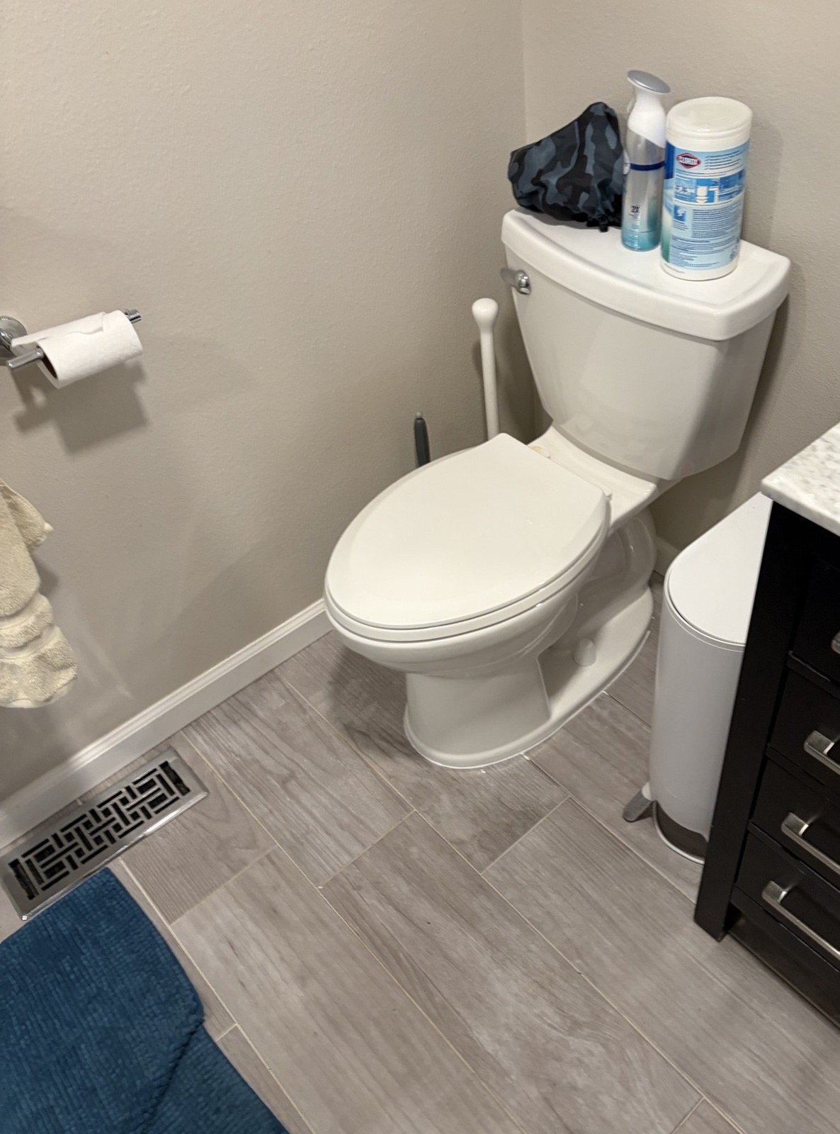 A white two-piece toilet back in place in an upstairs bathroom with grey plank tile, a dark vanity and a floor register beside it