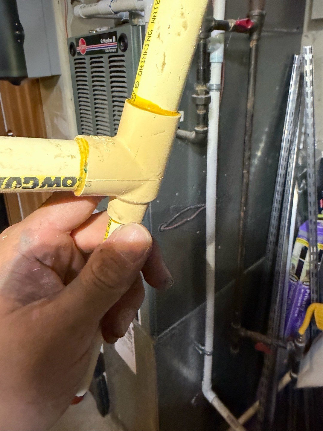 A plumber's hand holding up a yellowed CPVC tee with a hairline crack, pipes printed for drinking water, and a gas furnace behind it in a Duvall garage