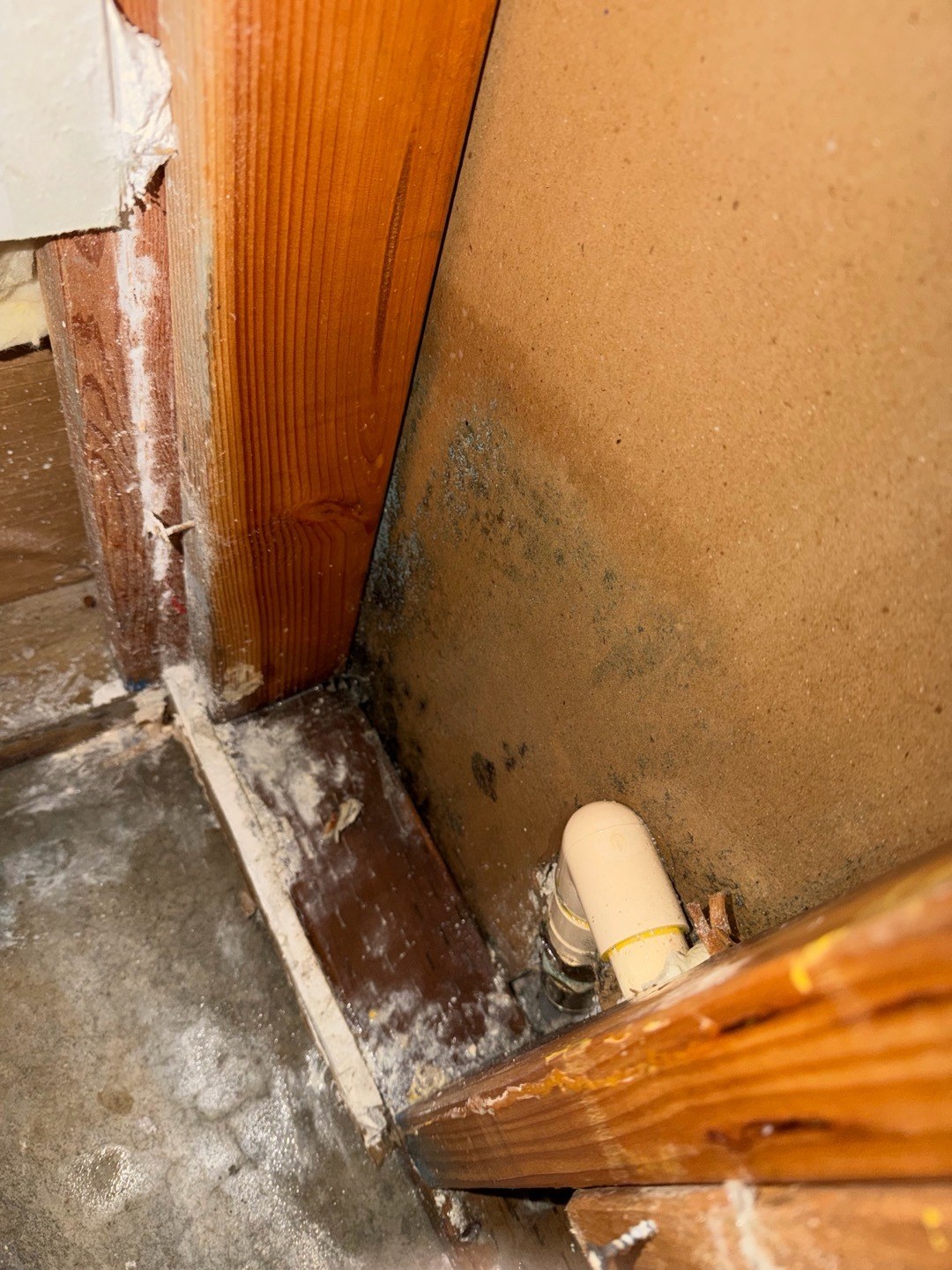 Open stud bay in a garage wall with a cream CPVC elbow rising through the bottom plate and dark water staining on the wall board above it