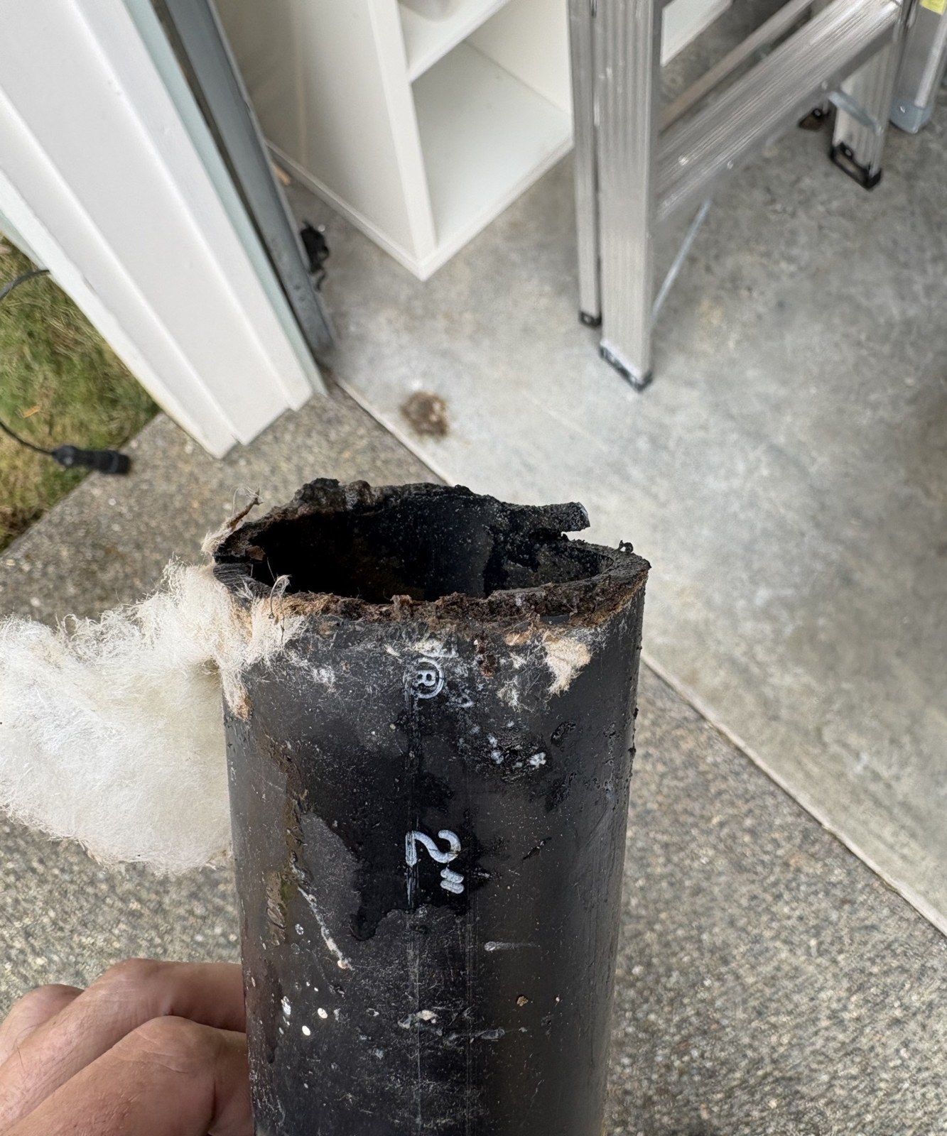 A hand holding a short length of old black two-inch ABS drain pipe with a ragged, broken end and insulation fibers clinging to it, a garage floor and a ladder behind