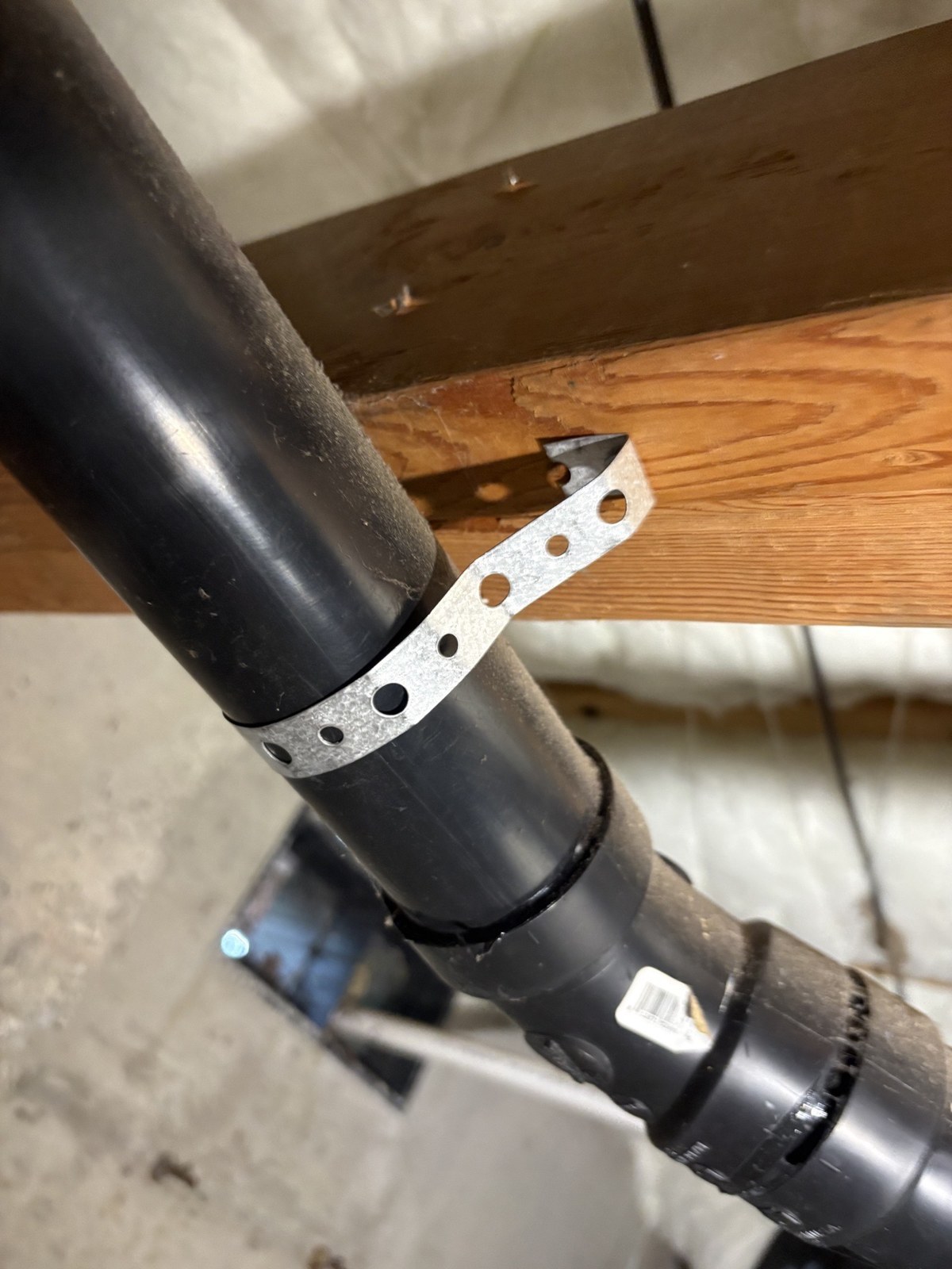 Black ABS drain pipe held to a wooden floor beam with a perforated metal hanger strap, with drain fittings below it in the crawlspace