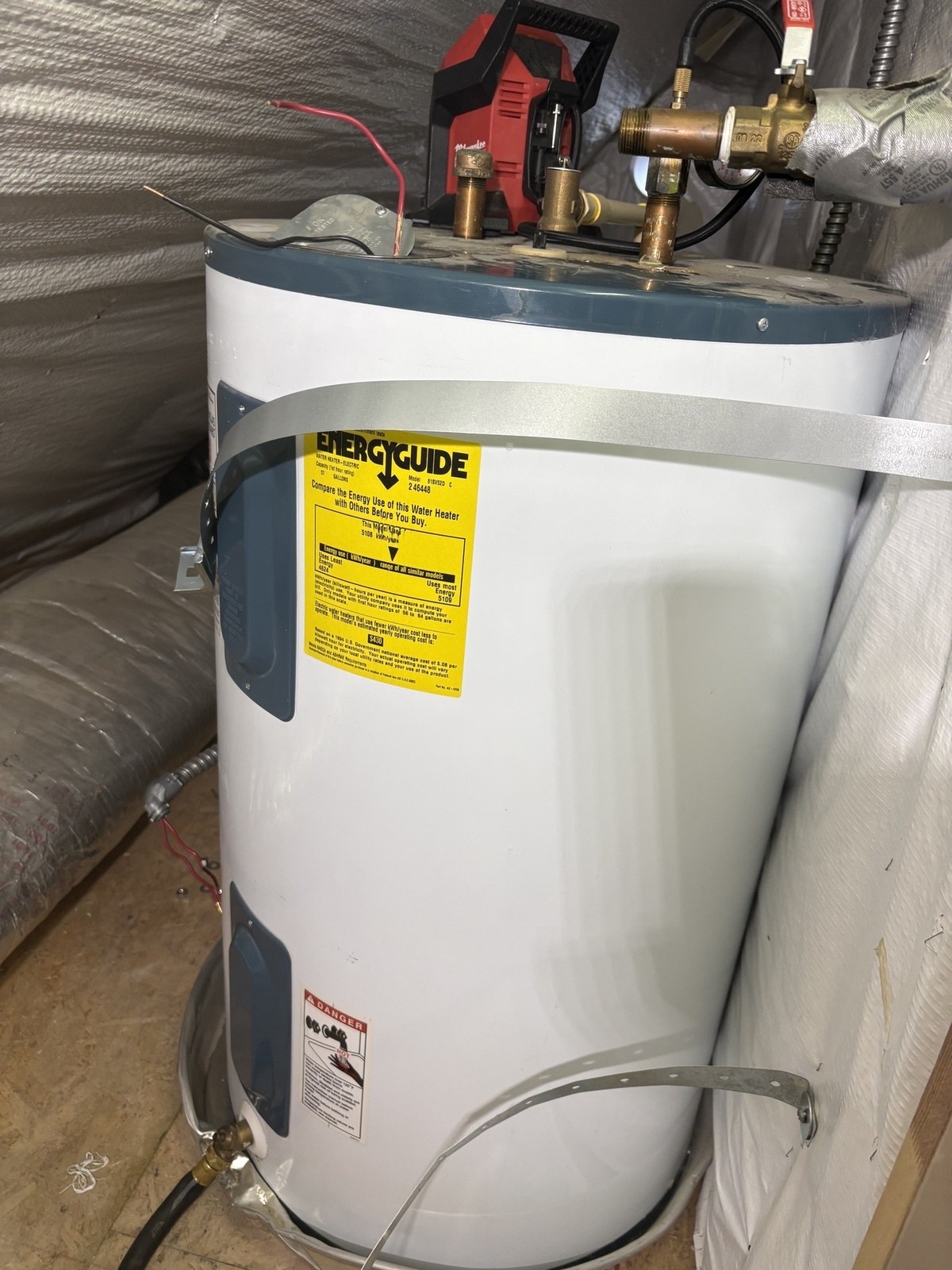 An old white electric water heater with a yellow EnergyGuide label in an attic space lined with insulation, a drain pump sitting on top while it empties