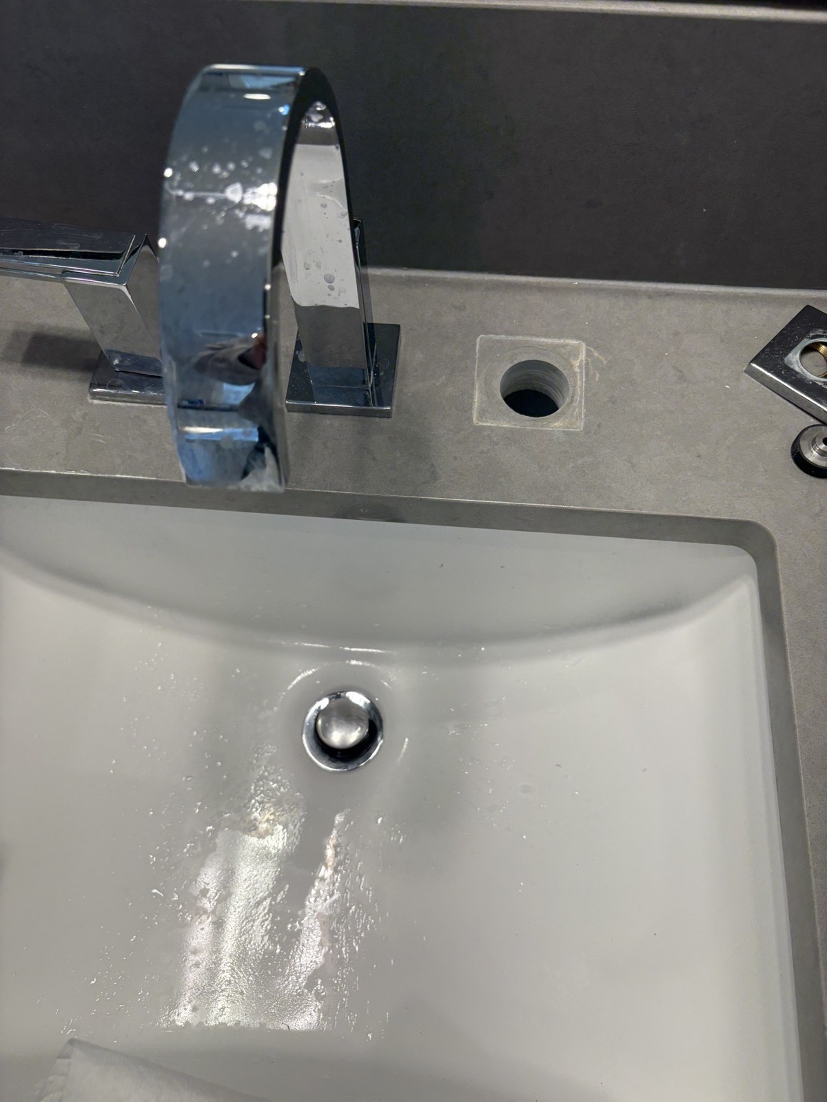 A chrome widespread bathroom faucet on a grey quartz counter with one lever handle missing, leaving an open valve hole beside the arched spout over a white undermount sink
