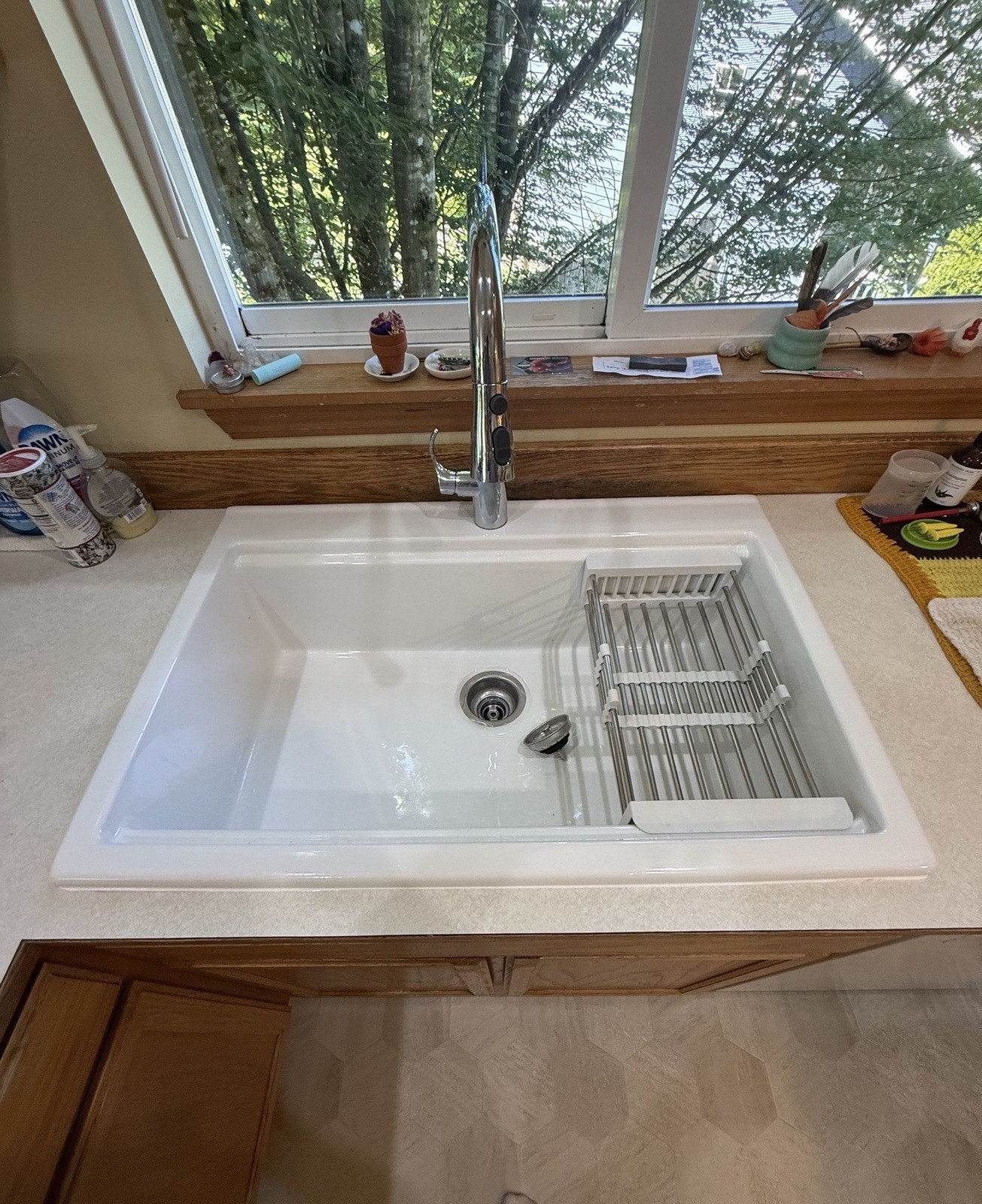 A white single-bowl cast-iron sink with a tall chrome pull-down faucet under a kitchen window, a dish rack in the bowl and trees outside