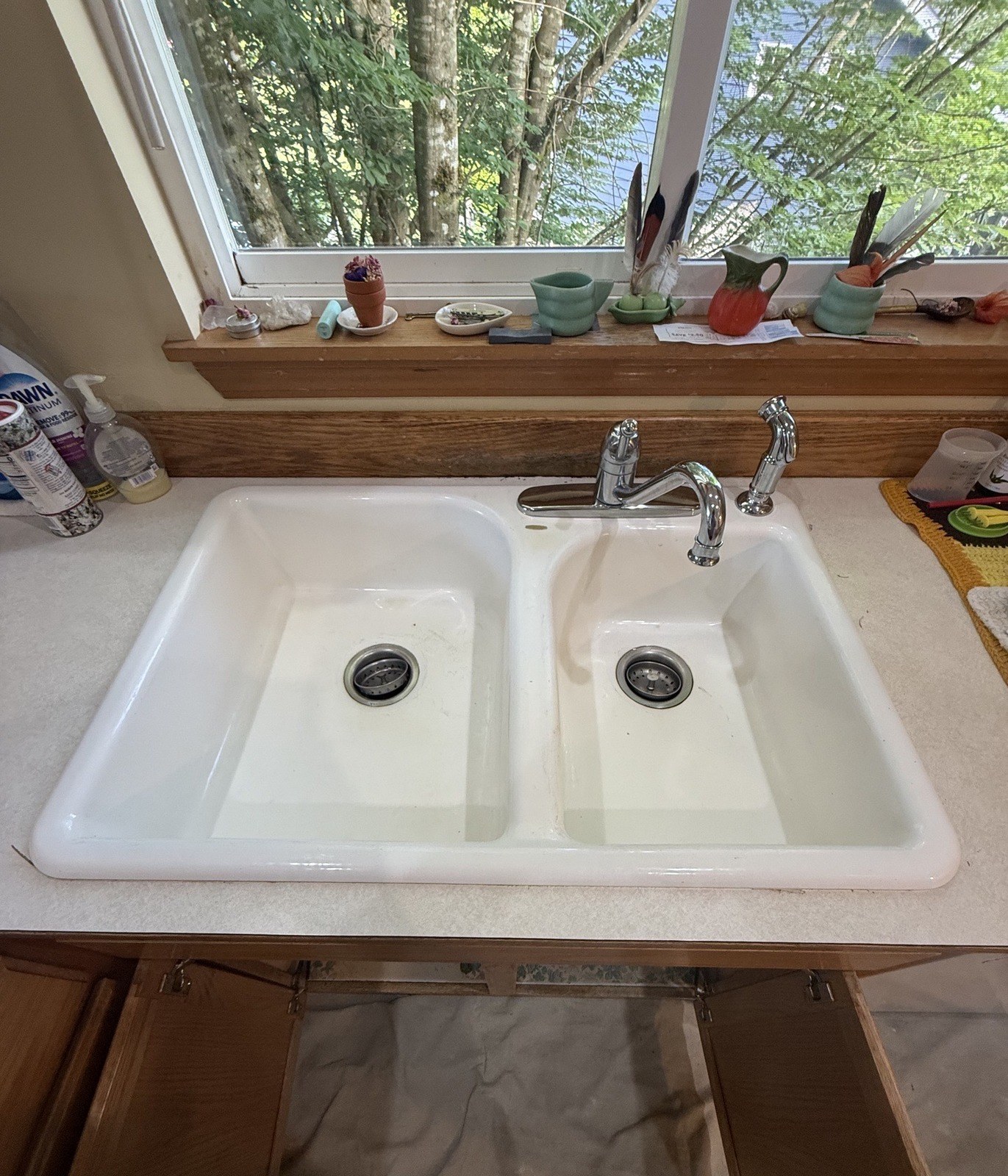 A worn white double-bowl kitchen sink under a window, with a chrome single-lever faucet, a separate side sprayer and the cabinet doors open below