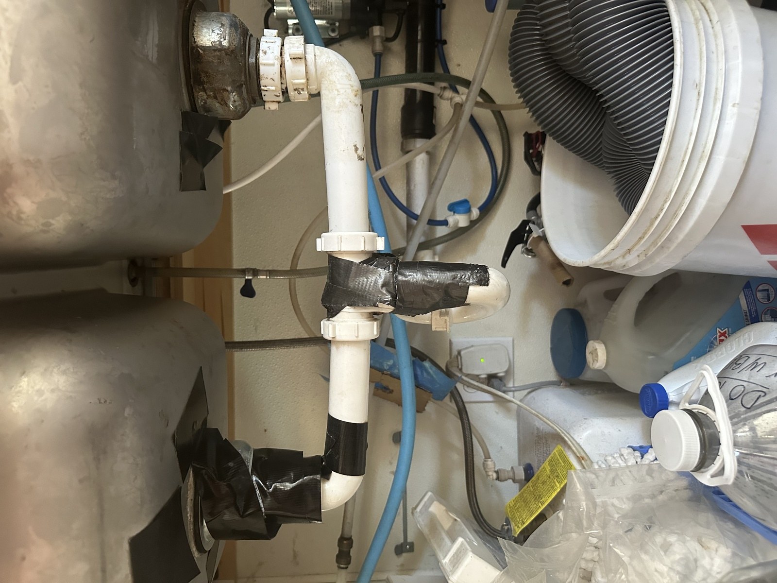 Cluttered under-sink cabinet at a coffee stand with rusted basket strainers and drain joints wrapped in tape