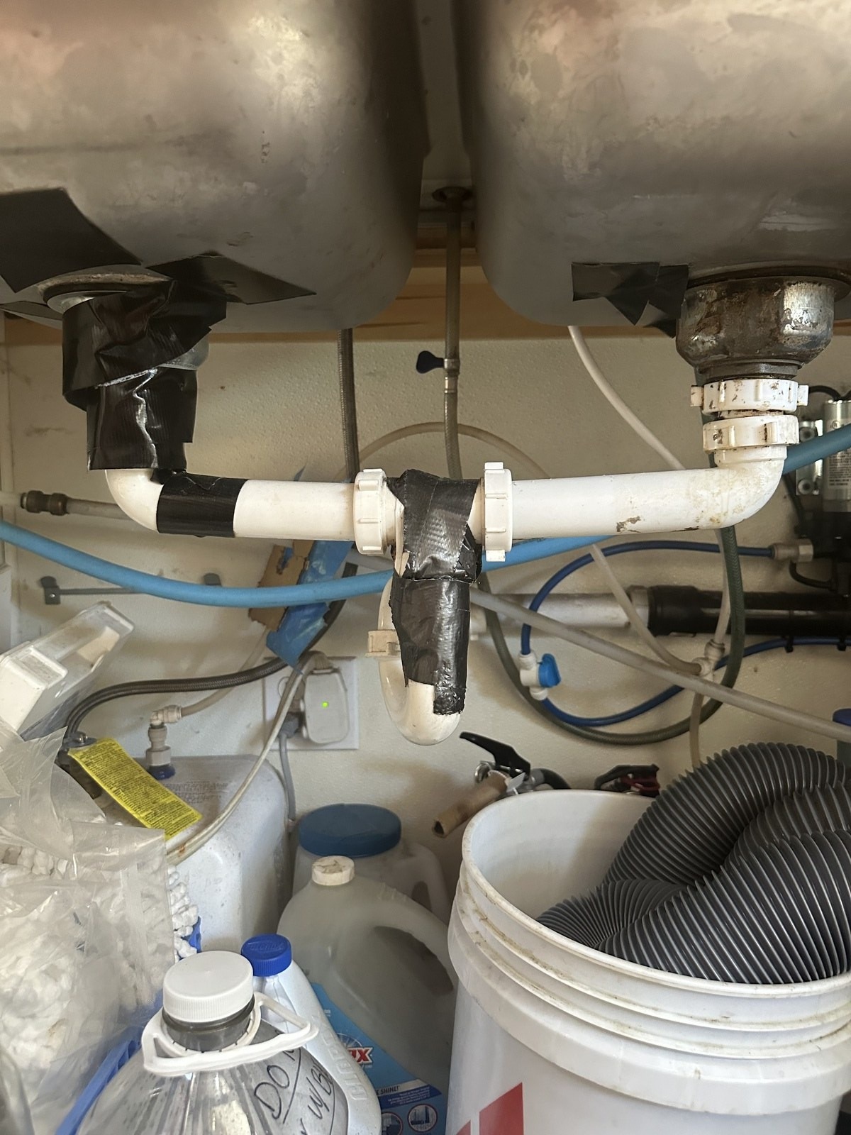 Cluttered under-sink cabinet at a coffee stand with rusted basket strainers and drain joints wrapped in tape