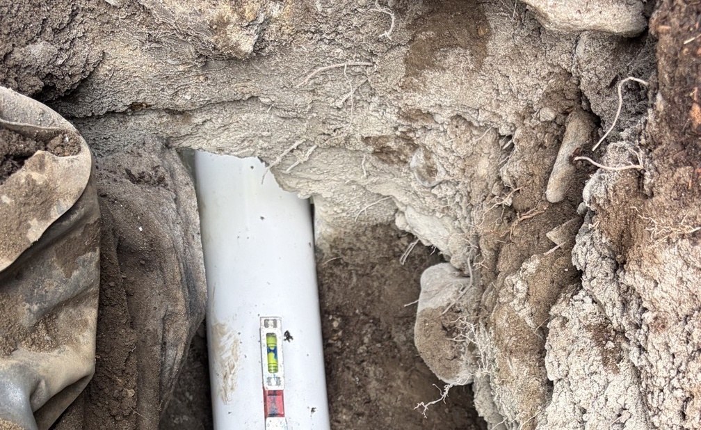 A new white 4-inch PVC sewer pipe lying on graded soil in a fresh excavation with a torpedo level resting on top of it, river rock and roots in the trench walls