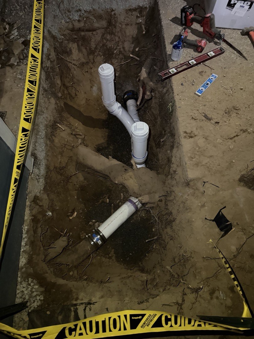 An open trench beside a house at night with a new white PVC double cleanout rising from the sewer line, a level, a primer can and a drill on the dirt beside it and caution tape around the edge