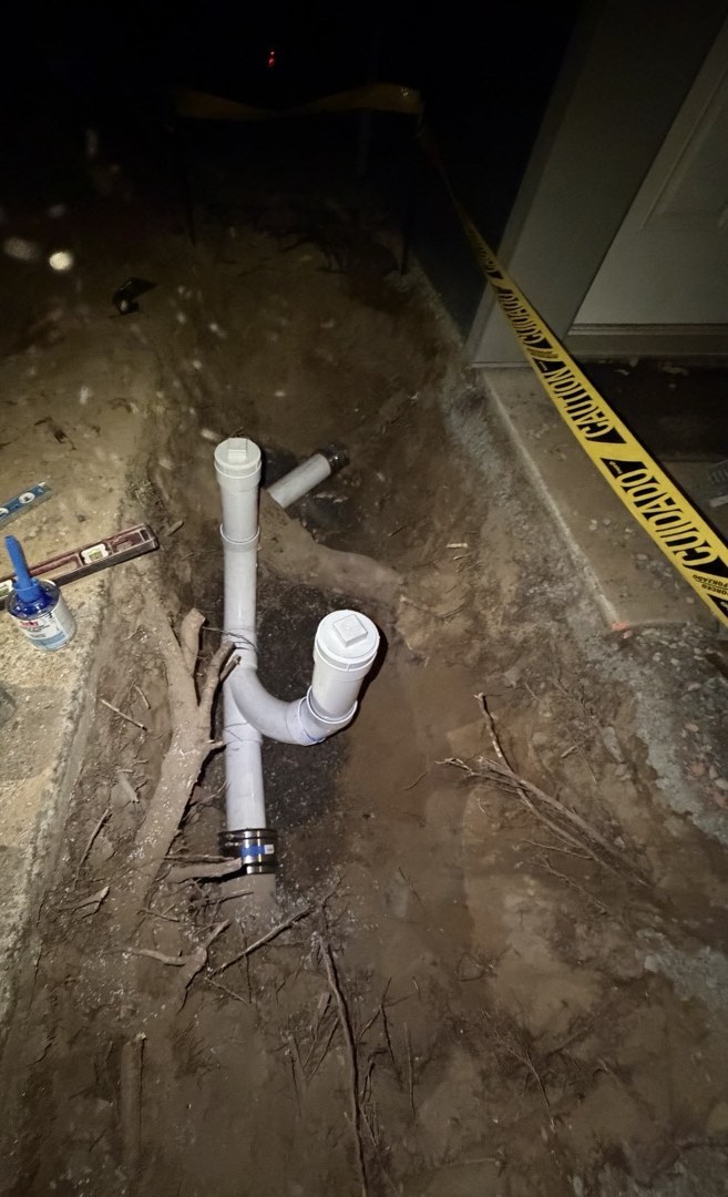 A narrow trench at night where a white PVC sewer pipe with two capped cleanout risers joins the old line, tree roots running through the soil and tools laid on the ground beside it
