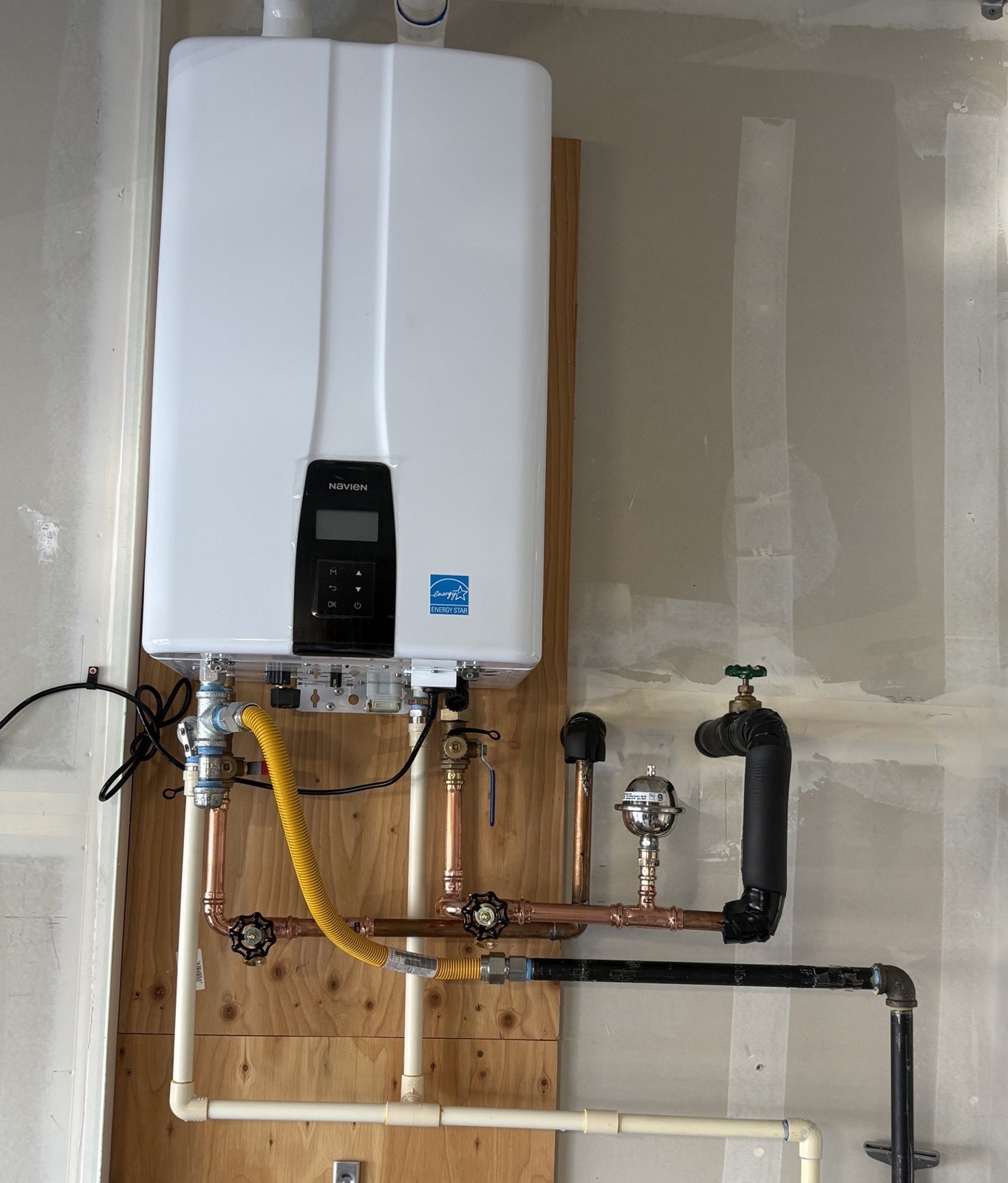 New white Navien NPE-240A2 tankless water heater mounted on a plywood backer, with new copper piping, a yellow gas flex, isolation valves and black insulated lines beneath it