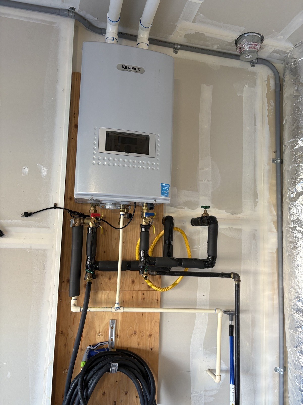 Old white Noritz tankless water heater on a plywood backer in a garage, with PVC vent pipes above it and insulated copper lines, a yellow gas flex and a coiled hose below